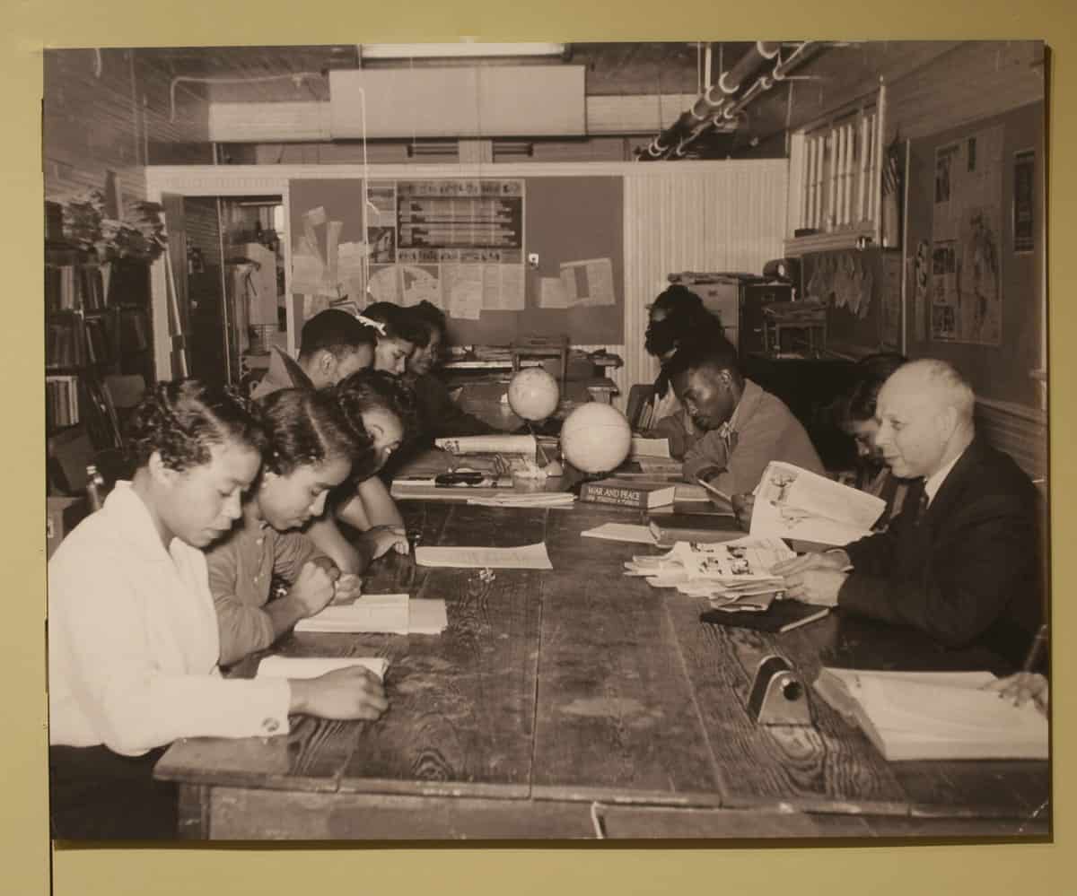 A historical image of a group of people sitting around a long table studying and doing work together. There are black people and white people together.